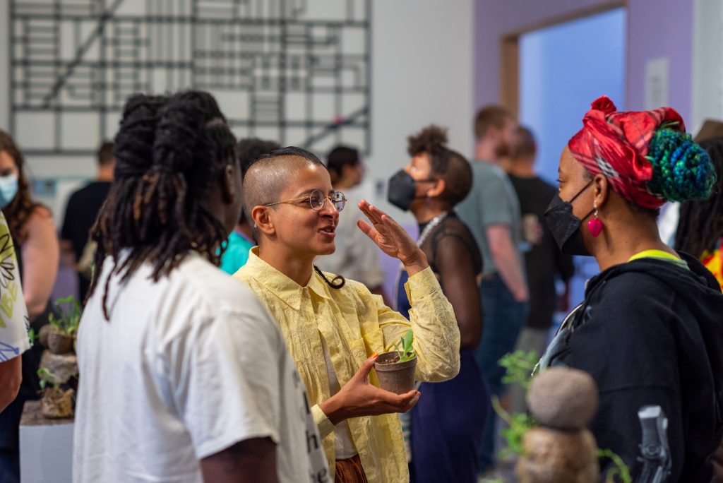 Image: Attendants at piecemeal opening night. In the foreground is a person facing away from the camera towards a a speaker wearing a yellow linen collared shirt and holding a plant gesturing with their free hand and smiling. They speak to a person wearing a black mask, a red head scarf and a pink earring. Behind them are crowds of people. On the back wall of the space is series of rectangular thick black lines. Photo by Tonal Simmons. 