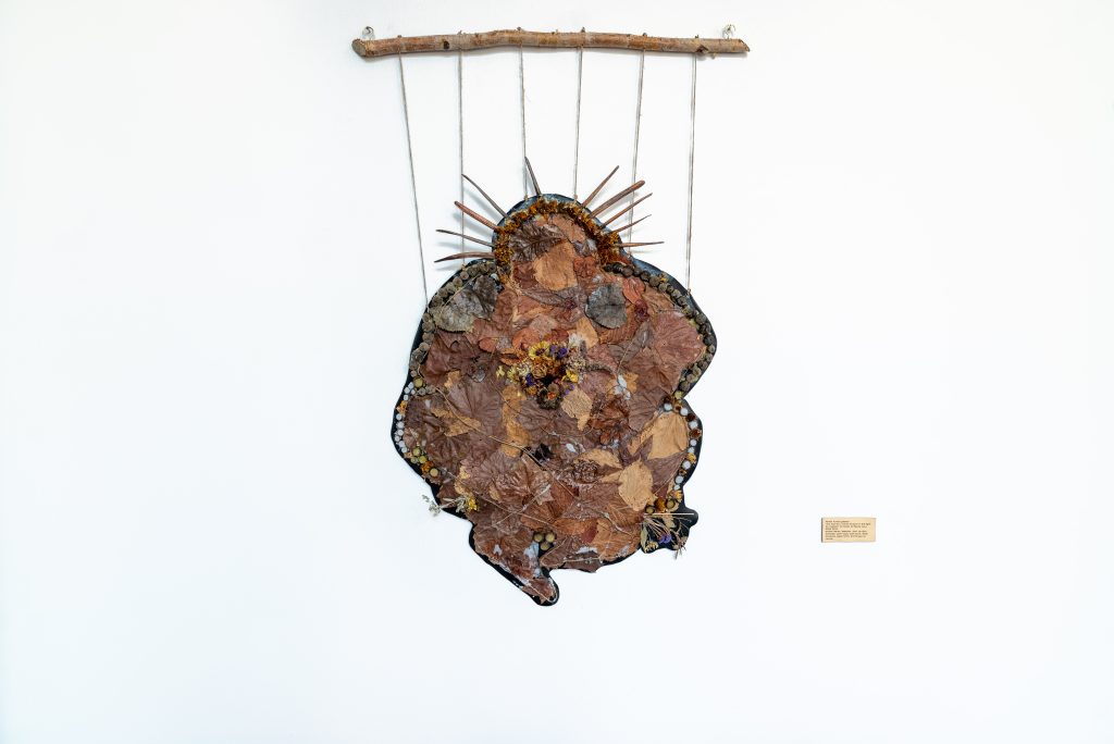 Image: darien golston, five foot four inches forward in the fight for freedom. An organic and irregular silhouette coated with dried leaves, flowers, and outlined by buttons, and acorn tops with a crown of sticks pointing out from its top.  It is connected and hung from a stick by six pieces of string. Photo by Tonal Simmons.