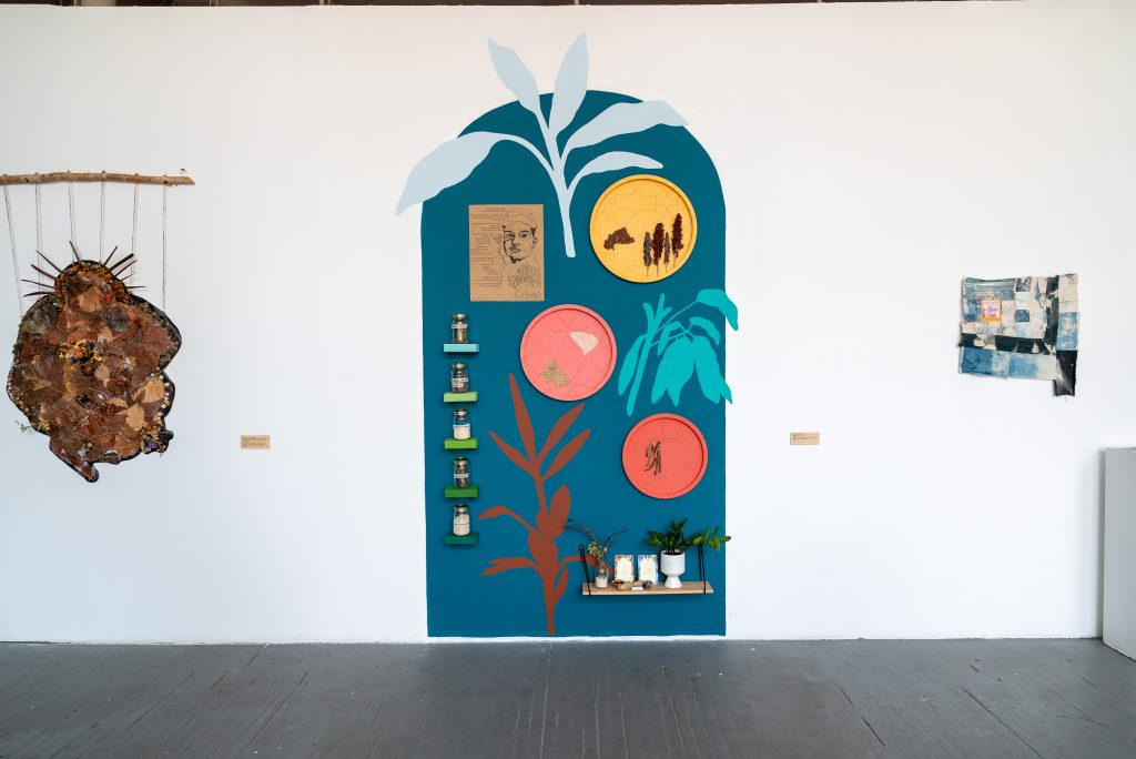 Image: Installation shot, alexandra antoine, seeds that bore fruit. A gallery wall, at the center of which is a blue archway painted on the door. On the door, are painted silhouettes of plants in teal, grey-blue, and maroon. There are several shelves, one on the bottom right, which has several vessels with plants and cards, and a column of five on the left side, each of which has a jar. There are three plates—yellow, pink, and salmon-colored—hung in the archway. There is a brown piece of paper with a drawing of a person's face in the top lefthand of the arch. Photo by Tonal Simmons.