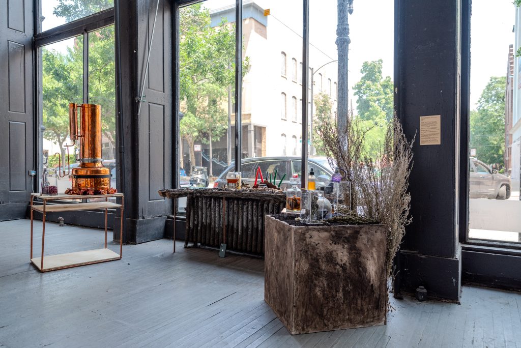 Image: Forrest Parks, Alchemical Memories: Ritual & Scent Mapping. In front of three tall, street-facing windows, three tables made of copper and wood, have various accoutrement on them. The left has a tall copper distiller. The middle one has several candles and glass vessels. The right one has vessels  and branches standing behind it. Photo by Tonal Simmons.