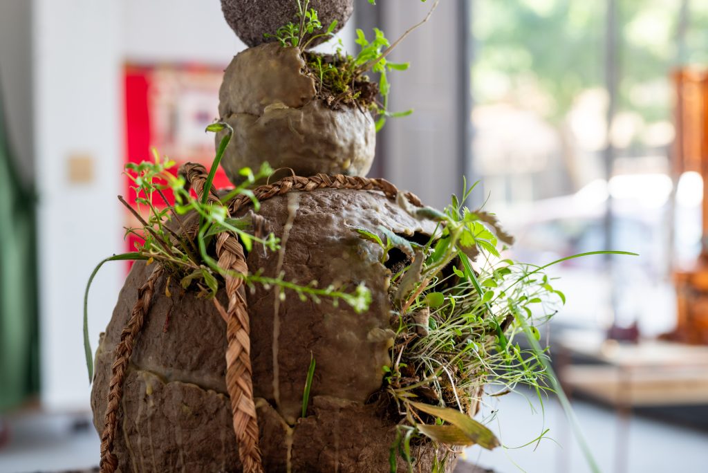 Image: Closeup, darien golston, Earthen Vessel #1. A stack of three clay orbs, smaller as the stack rises, each rupturing out with dirt and sprouts. The bottom one is encircled with a rope. In the background, out of focus, a day lit room. Photo by Tonal Simmons.