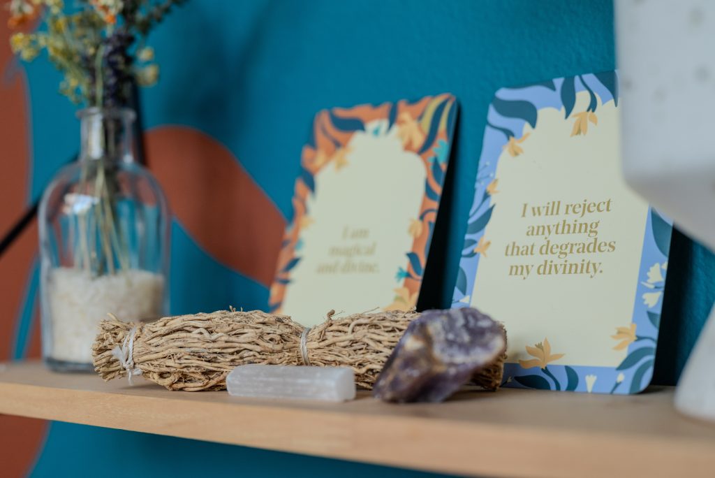 Image: Close-up of seeds that bore fruit. A shelf with an herb bundle, crystals, and a bottle filled with dried flowers. There are two cards up against the wall, one outlined blue, the other outlined orange. In the center of each there is affirmational text: "I will reject anything that degrades my divinity." and "I am magical and divine." Photo by Tonal Simmons.