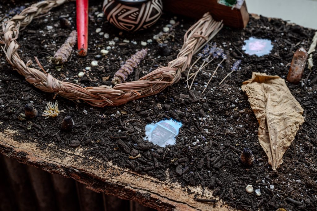 Image: Forrest Parks, Alchemical Memories: Ritual & Scent Mapping.A surface covered with dirt and wood chips. Embedded or laid upon the dirt are dried flowers, dried leaves, woven ropes, a red candle.  Spots where the dirt is cleared away reveals photos of faces. Photo by Tonal Simmons.