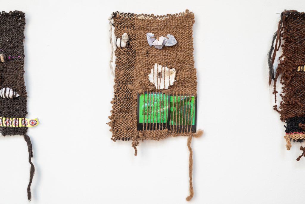 Image: Bryana Bibbs, Journal Series.  Three woven collages. The center one is made with a beige burlap-like material. Woven into it are four stones and a lime green panel in the bottom right hand corner. A strand of the weaving material hangs down from the bottom right hand corner. The left and right ones  are made of a darker weaving material. Photo by Tonal Simmons.