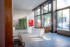 Image: Installation view, piecemeal: (earthen works in progress). The front room of Chicago Art Department in the midday. Three floor to ceiling windows let in a soft morning light cast across several art pieces. There is a green curtain to the left, a red quilt to the right of it, and before that three tables, one with a copper distiller on it. Photo by Tonal Simmons.