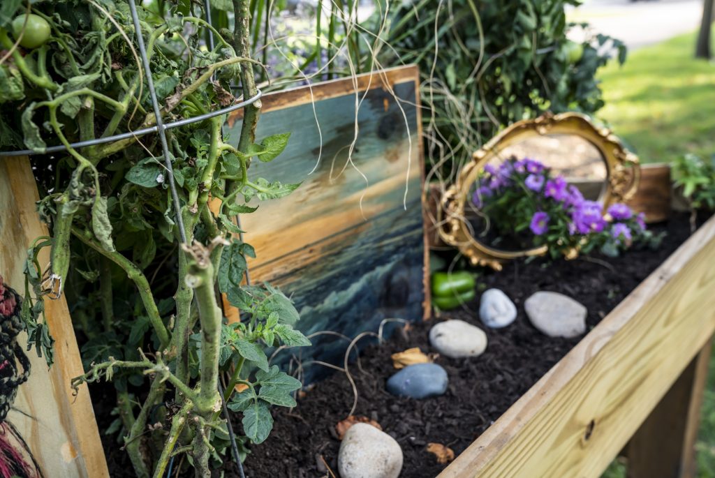 Image: Tending to My Garden, 2022 by Edna M. Togba, installed as part of Finding Ceremony—Tiffany Johnson's project for her 2022 Anchor Curatorial Residency at Austin Town Hall. This close up view shows a landscape painting placed among rocks, flowers, and a mirror in the earth. Image courtesy of Tiffany Johnson. Photographer: Vanessa Lara/ Colectivo Multipolar.