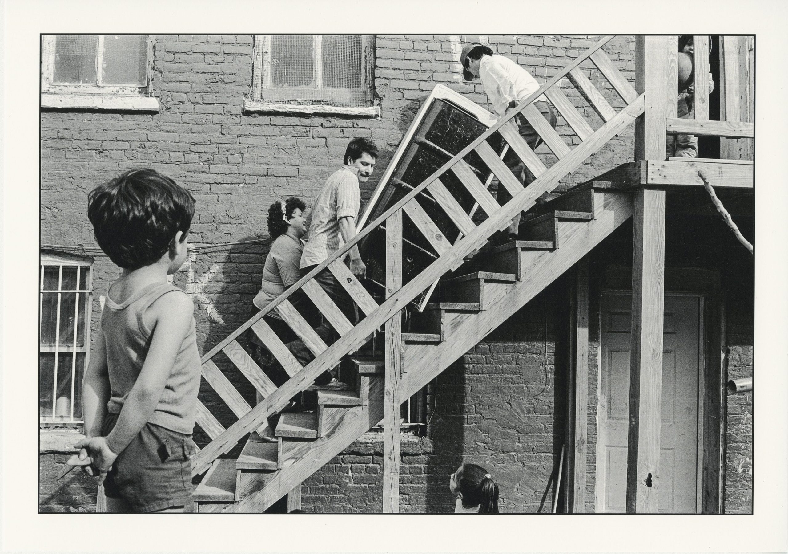 Photographer: Akito Tsuda. Chicago Public Library, Special Collections, Akito Tsuda Photographs, Box 1: Photograph 1.45. In this black and white photo, a boy and a girl watch two men and a woman moving an appliance up the wooden stairs behind an apartment.