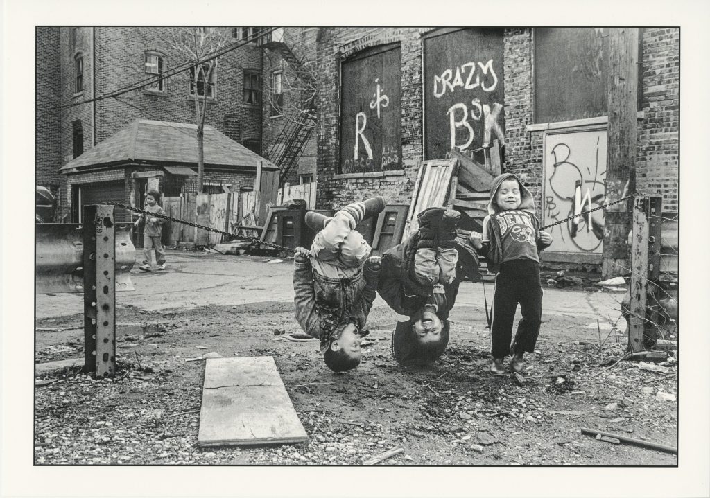 Image: Pilsen, circa 1991-1994, Photographer: Akito Tsuda. Chicago Public Library, Special Collections, Akito Tsuda Photographs, Box 1: Photograph 1.57. In this black and white photograph, three kids hang out at a DIY playground, two hanging upside down, giggling. 