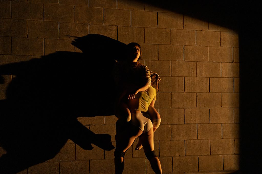 Image: Two figures are in a stark yellow light, against a painted wall with a grid of concrete bricks. One holds another against the wall, straddled by the lifted figure. The lifted figure's arm falls across the first's back. Their shadows are long and distorted against the wall. Photo by Michelle Reid.