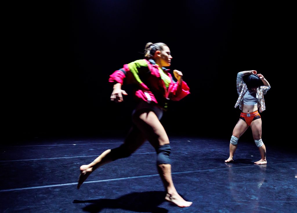 Image: On a scuffed black marley floor and under a harsh white light, a figure is mid-stride, blurred by their movement, in the foreground. They wear a knee brace and neon pink and green jacket. To their right, in the mid ground, another figure: both feet firmly on the ground, knees slightly bent and a little further than hip-distance apart. They wear a speedo and an unbuttoned plaid shirt over a white crop top. Their arms are raised over their head and their head turned to their side. Photo by Sarah Larson.