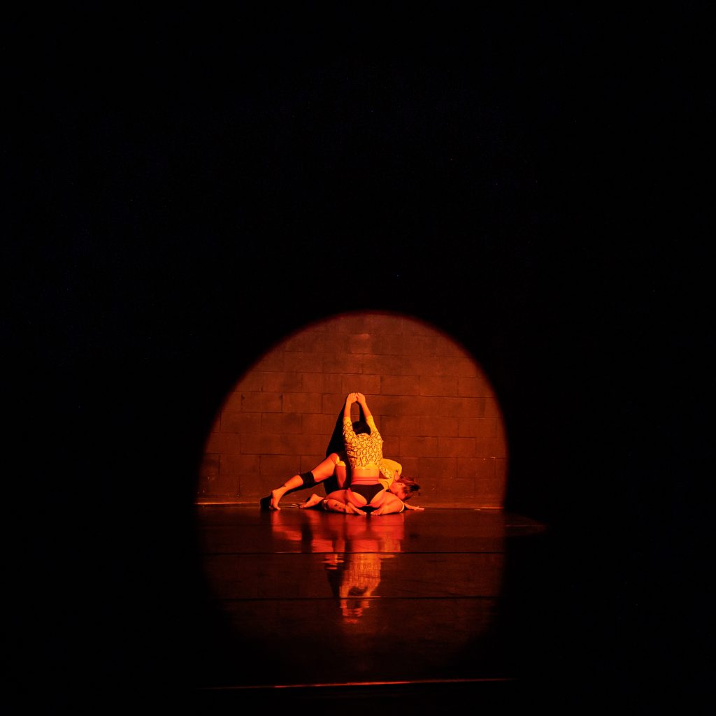 Image: In an orange spotlight, two dancers are near the ground against a wall. The dancer in front of the other kneels with their legs spread. The one behind is stretched out parallel to the floor, with one leg pressing its ball to the floor and their heel back. Their face is obscured by the other person's body. Surrounding the spotlight is darkness. The dancers are reflected on the floor. Photo by Sarah Larson.