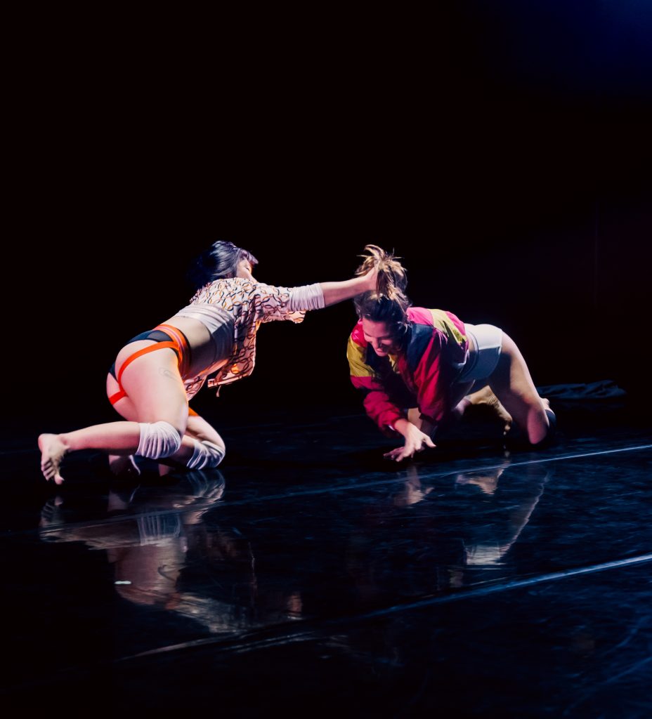 Image: Two figures are on a reflective black floor. The one on the left is on their knees, reaching over to the other, pulling their ponytail. The one on the right wears a neon pink and yellow jacket, crossing their hand in motion. Photo by Sarah Larson.