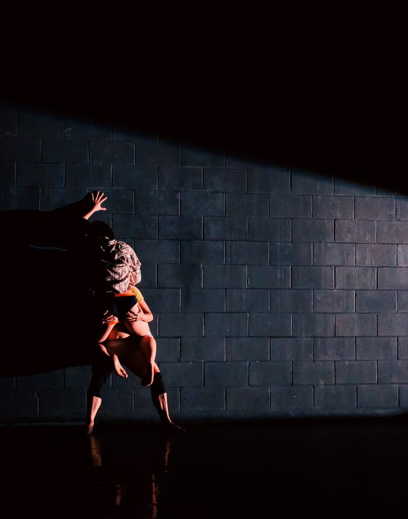 Image: A painted black wall, gridded concrete blocks. From the right, a stark white light is cast , leaving the two figures on the white in shadows. One figure stands , their legs visibly straining in touching the ground. Apart from their legs, only their hands are visible against the back of thighs, holding another figure in straddle against them. The second figure's left hand is against the wall. Neither's face is visible. Photo by Sarah Larson.