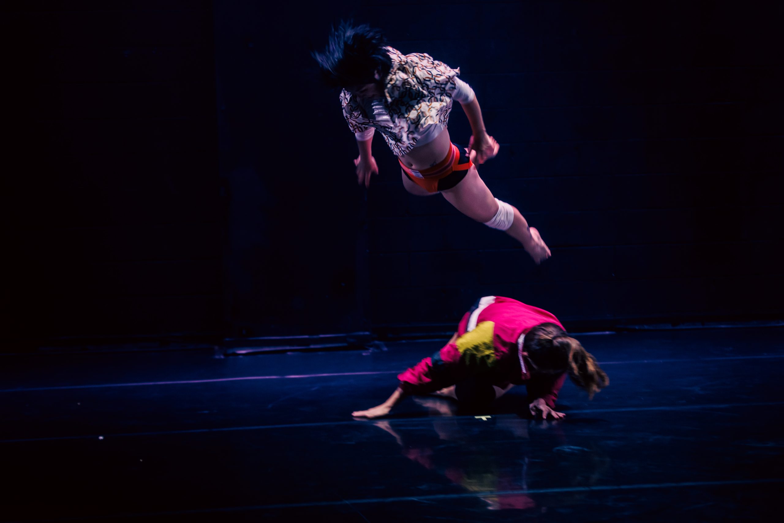 Image: Still from "Knockout." Two figures in a dark space. One, sweeps close to the ground, wearing a neon pink and yellow jacket. The other is nearly horizontal above them, hair sweeping with motion. Photo by Sarah Larson.