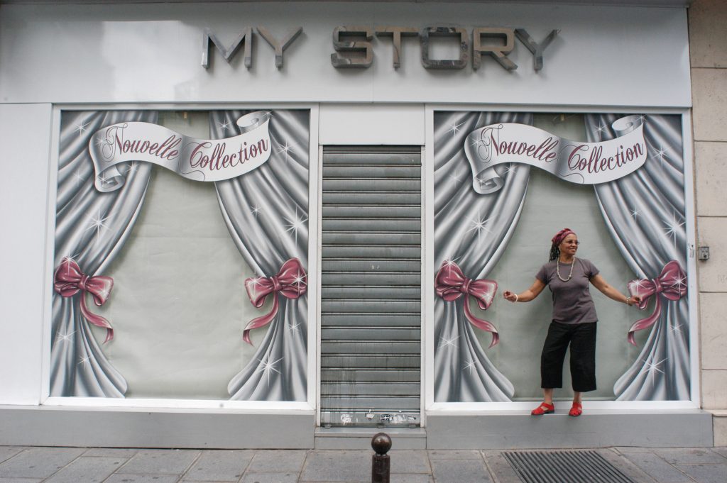 Image: A woman in red shoes poses against street art created to resemble an open shop window. Painted pink ribbons tie back the painted curtains and above the woman's head reads the words "Nouvelle Collection" in flowing script. Beverly Cottman in Paris Photo courtesy of writer. 