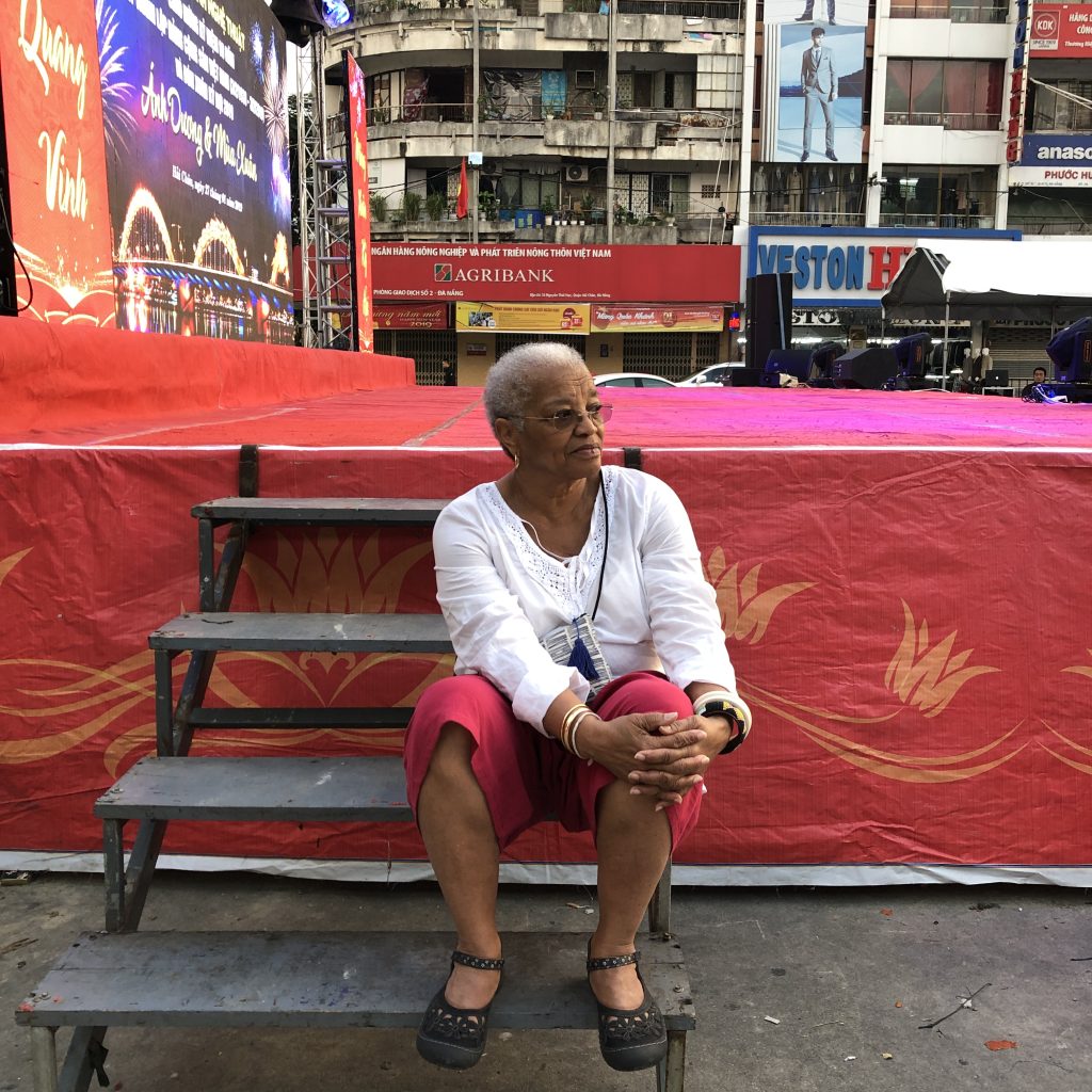 Image: A woman in a white blouse sits on wooden steps that lead to a colorful public stage/platform. Behind her and the stage there are a variety of city buildings and ads. Beverly Cottman in Da Nang. Photo courtesy of writer.