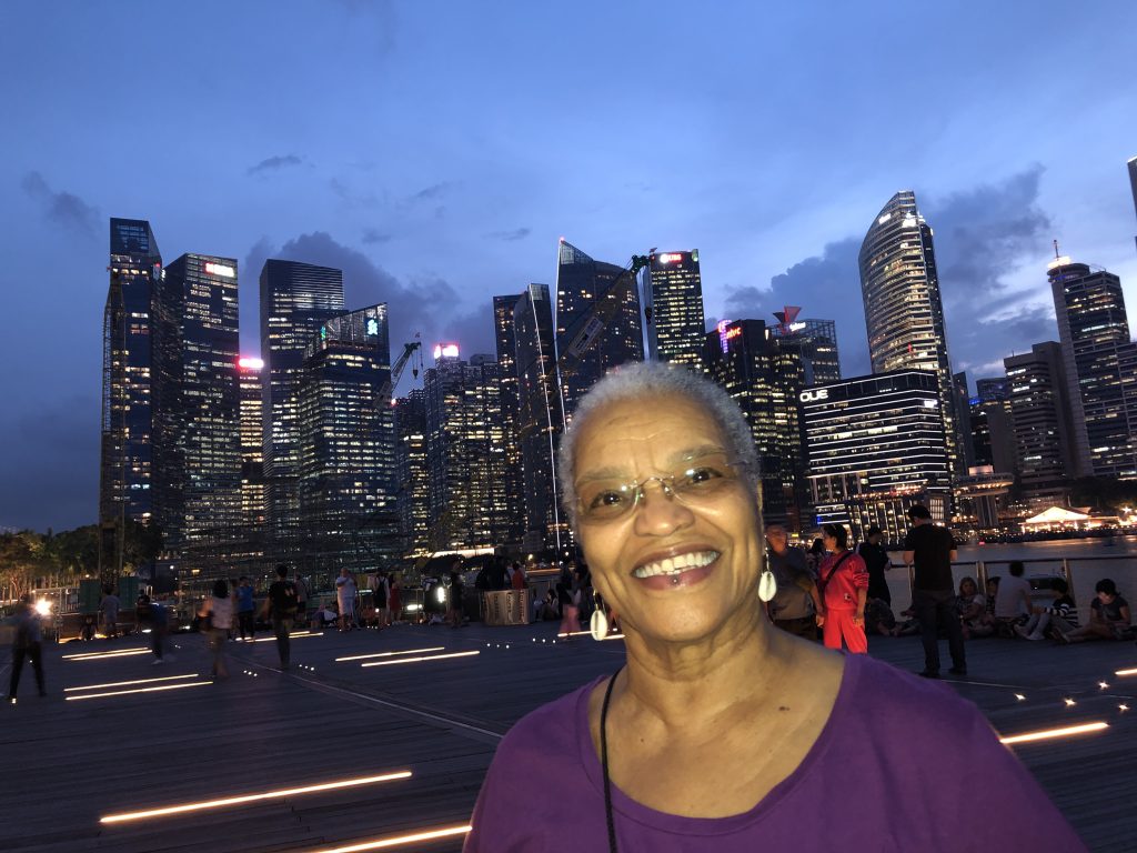 Image: A woman in purple wearing cowrie shell earrings smiles against a cityscape. Beverly Cottman in Singapore. Photo courtesy of writer. 