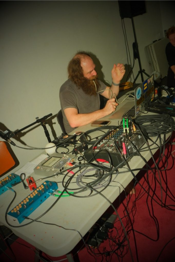 Image: Ryan Dunn performs his sound project, maoϴ. Dunn sits at a plastic folding table covered with sound equipment. A plastic tube runs from his equipment to his mouth. Photo by Michael Wellvang. Image courtesy of Anna Johnson.