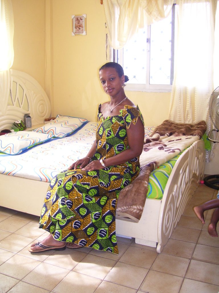 Image: A woman in a dress made from traditional African textiles sits on a bed in a sunlit room. Kenna Cottman in Banjul. Photo courtesy of writer.