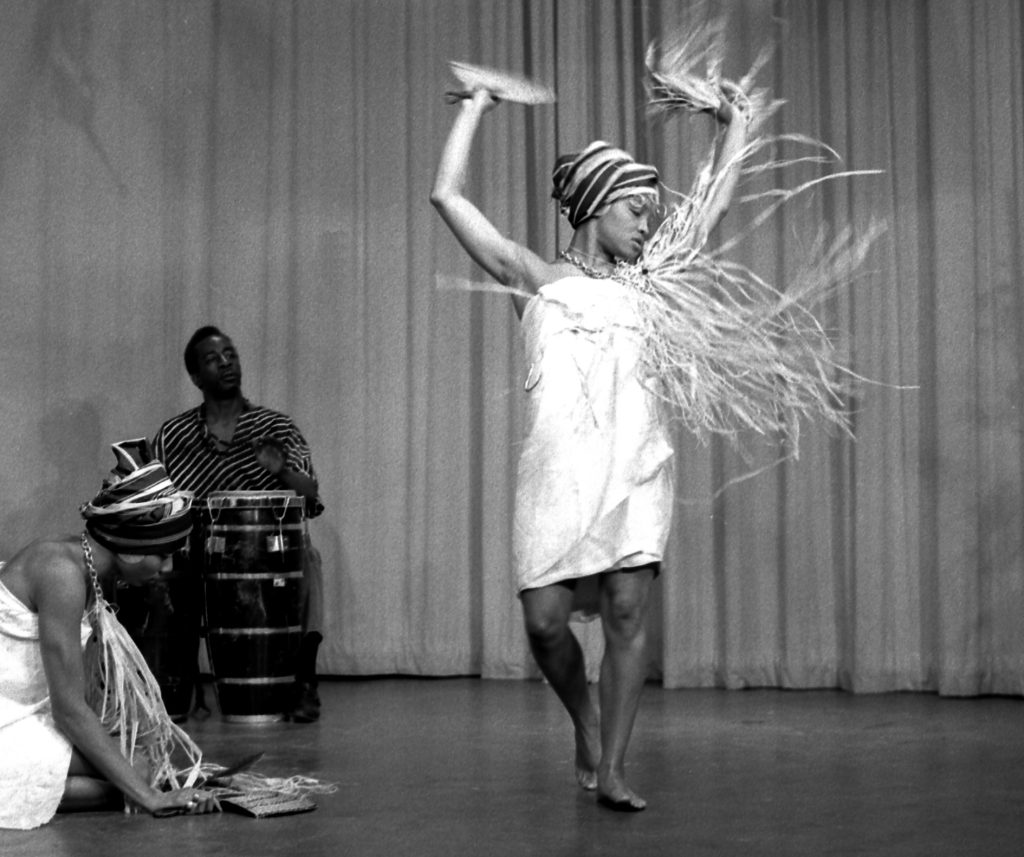 Image: A woman dances on stage in a white dress, her hands are raised mid-movement. A man sits behind her with drums and another woman in white sits on stage to her side. Beverly Cottman performs with a dance company. Photo courtesy of writer.