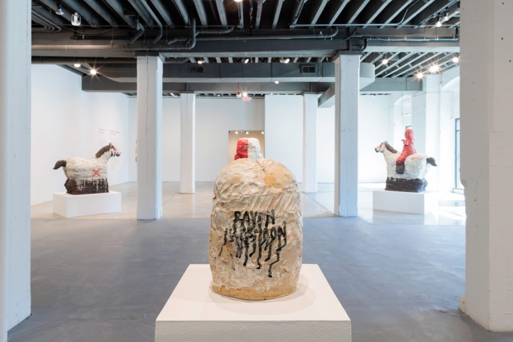 Image: Installation of Raven Halfmoon: Flags of Our Mothers, 2024. On view at Bemis Center for Contemporary Arts. Courtesy of Bemis Center for Contemporary Arts. A view of a large gallery space with several large-scale sculptures, which are mostly black, red, and white. The sculpture in the foreground is a round, organic shape and sits on a pedestal. It is off-white in color and has the name “RAVEN HALFMOON” abstractly written in black in the center. Photography by Colin Conces. 