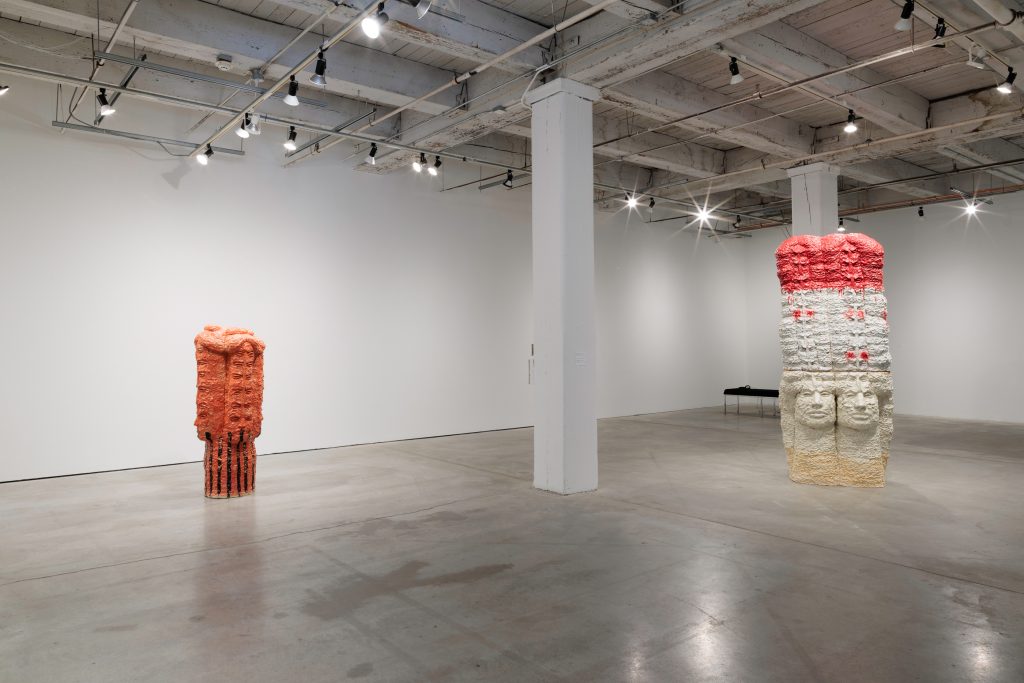 Image: Installation of Raven Halfmoon: Flags of Our Mothers, 2024. On view at Bemis Center for Contemporary Arts, May 18-September 15, 2024. A view of a large gallery space with two sculptures: the sculpture to the left is mostly red with some black and depicts three faces looking in different directions. The top section of the faces are extended and repeated upwards. The sculpture on the right is very large and is mostly red, white, and off-white. It depicts two faces towards the bottom with sections of smaller faces repeated and extending upwards. Courtesy of Bemis Center for Contemporary Arts. Photography by Colin Conces.