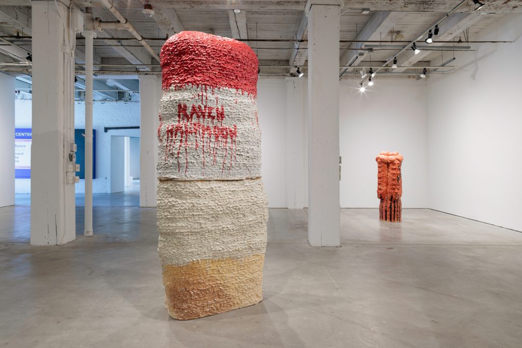 Image: Installation of Raven Halfmoon: Flags of Our Mothers, 2024. On view at Bemis Center for Contemporary Arts, May 18-September 15, 2024. A view of a large gallery space with two sculptures: the sculpture to the left is very large and is mostly red, white, and off-white. The name "RAVEN HALFMOON" can be seen in red on the sculpture dripping down. The sculpture on the right is much smaller and is mostly red with some black and depicts three faces looking in different directions. The top section of the faces are extended and repeated upwards. Courtesy of Bemis Center for Contemporary Arts. Photography by Colin Conces.