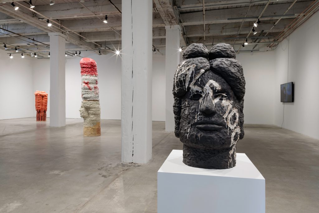 Image: Installation of Raven Halfmoon: Flags of Our Mothers, 2024. On view at Bemis Center for Contemporary Arts. Courtesy of Bemis Center for Contemporary Arts. A view of a gallery space with several large-scale sculptures on display. The sculpture in the foreground depicts a large head sitting on pedestal. The sculpture is all black save for the name “HALFMOON” written across it in white. The sculptures in the background are mostley red, white, off-white, and black. Photography by Colin Conces. 