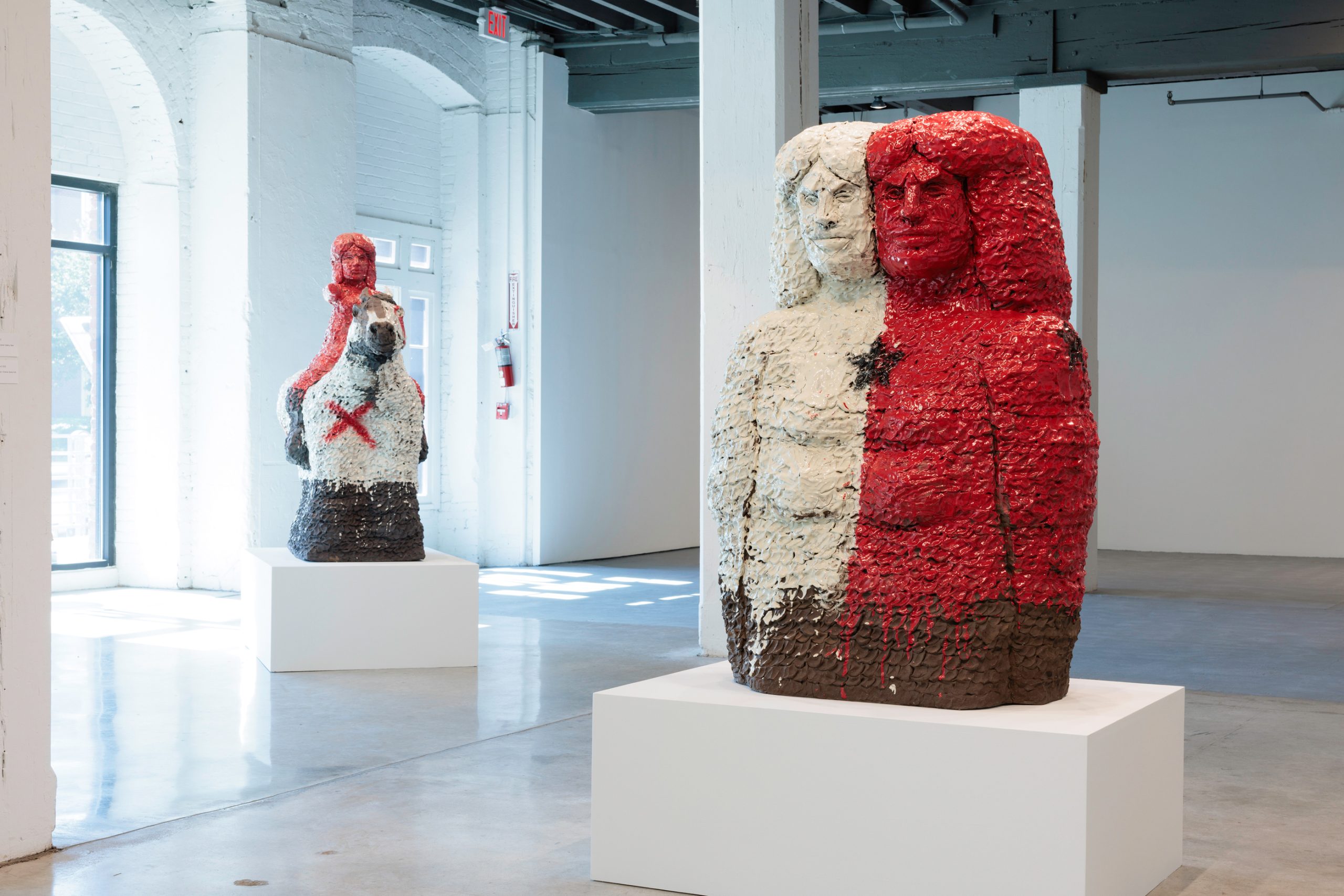 Image: Installation of Raven Halfmoon: Flags of Our Mothers, 2024. On view at Bemis Center for Contemporary Arts. Courtesy of Bemis Center for Contemporary Arts. A view of a large gallery space with several large-scale sculptures, which are mostly black, red, and white. The sculpture on the right depicts two figures connected in the middle. The sculpture on the left depicts a woman on a horse: the woman is red and the horse is white. Photography by Colin Conces.