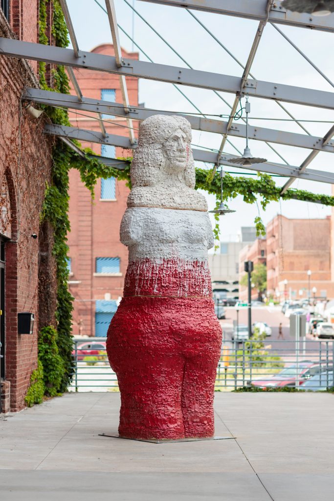 Image: Installation of Raven Halfmoon: Flags of Our Mothers, 2024. On view at Bemis Center for Contemporary Arts, May 18-September 15, 2024. Flagbearer by Raven Halfmoon can be seen on view outside the gallery space. It is 14-feet tall with a red base, a white middle that drips onto the red, and an off-white top. The sculpture depicts an Indigenous woman. Courtesy of Bemis Center for Contemporary Arts. Photography by Colin Conces.