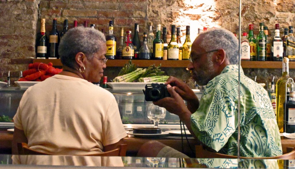 Image: A man and a woman sit at a bar, as the man shoes the woman a detail on the camera he holds. The brick wall behind them holds shelves of many colored liquors and there are plates of asparagus and peppers upon the lowest shelf. Beverly and William Cottman in Barcelona. Photo courtesy of writer.