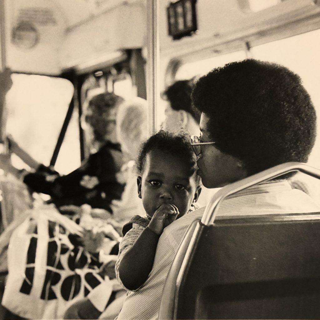 Image: A woman and a baby sit on a bus seat. The baby is turned to face the camera, the woman looks forward while holding the child. Beverly Cottman with Kenna Cottman. Photo courtesy of writer.