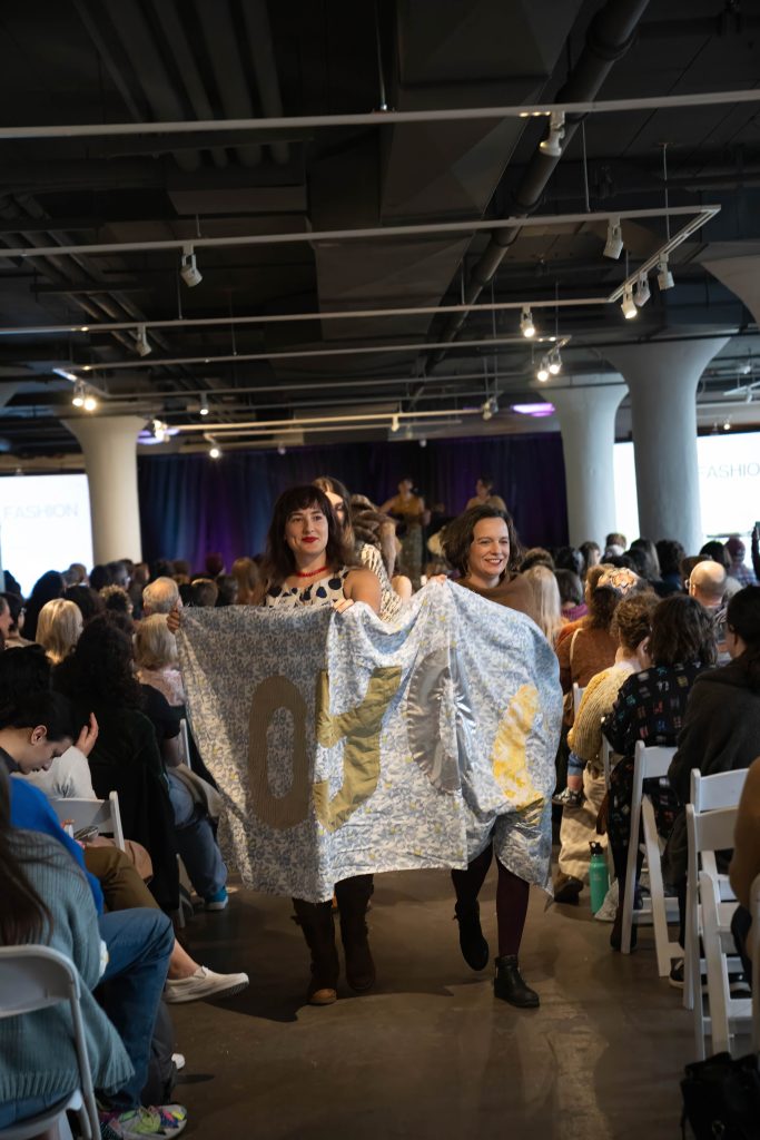 Image: People walking down a runway holding a quilt. Photo from the second Cohort of One Year One Outfit's fashion show at the 2024 Rust Belt Fibershed Symposium. Photo Courtesy of Michael Kent at Kogent Studios.