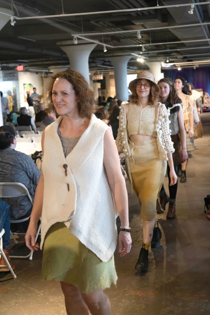 Image: People walking down a runway wearing outfits made from the second Cohort of One Year One Outfit's fashion show at the 2024 Rust Belt Fibershed Symposium. Photo Courtesy of Michael Kent at Kogent Studios.