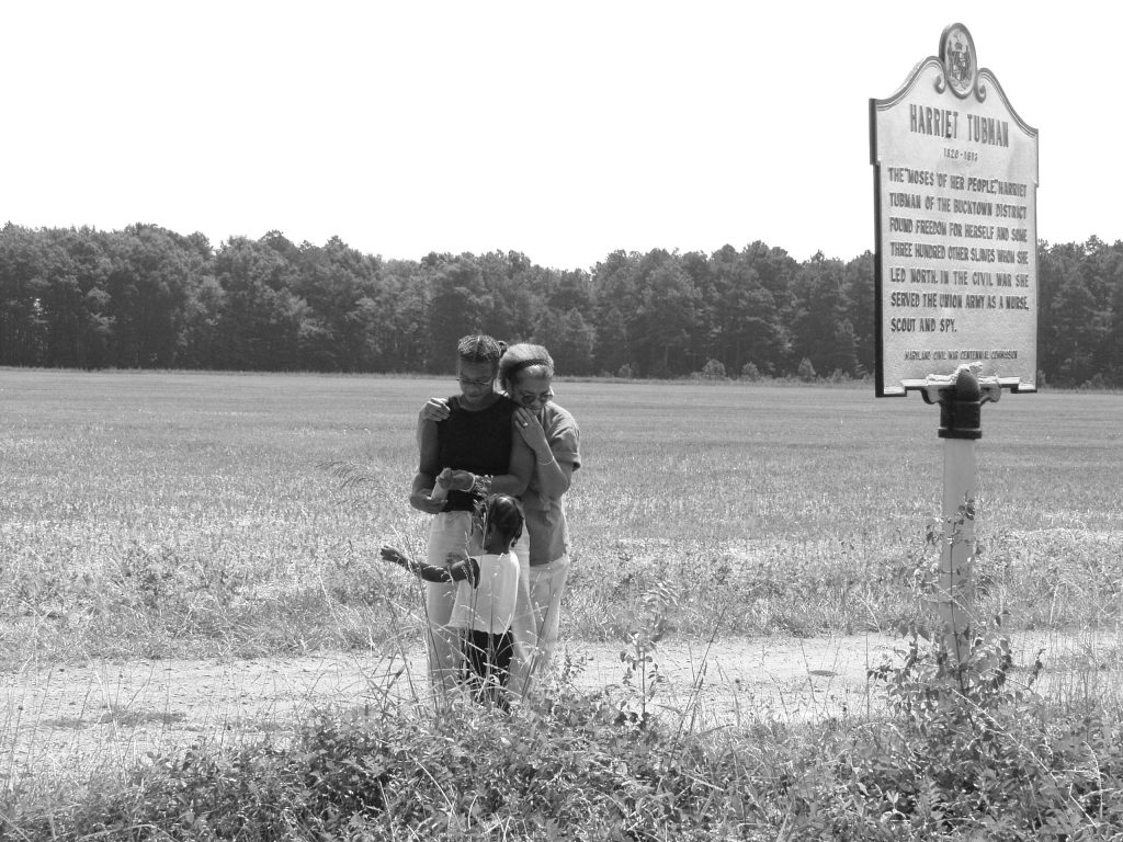 Image: Three women embrace in a sunny field near a cleared pathway. Kenna, Beverly Cottman, and Yonci Jameson at the Harriet Tubman Underground Railroad State Park in Maryland. Photo courtesy of writer.