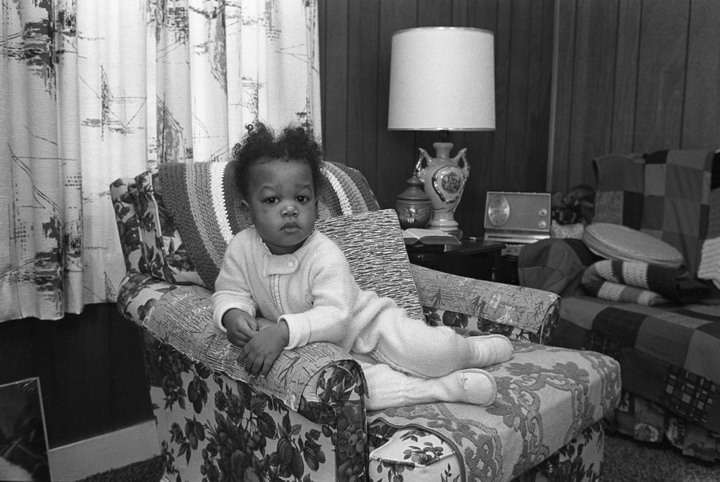 Image: In a black and white image a baby in a light colored onesie leans forward on a decorated couch. Behind her is a lamp and a window with light curtains captured mid-wave. Kenna Cottman, photographed by William Cottman. Photo courtesy of writer.