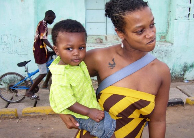 Image: A woman holding a young boy is caught mid-sentence. Behind them there is a vibrant blue wall against which a man walks with his bicycle. Kenna Cottman and a young Ebrima in Banjul. Photo courtesy of writer.