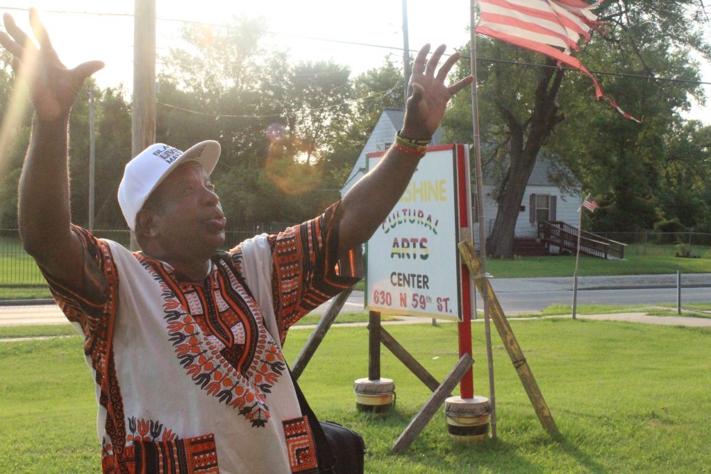 Image: Sylvester "Sunshine" Lee stands with his arms raised outside in the sun. Behind him stands a sign for his Cultural Arts Center, which also bears his name. The street behind him is empty, no other people are in frame, 2024. Photo by Treasure Shields Redmond.