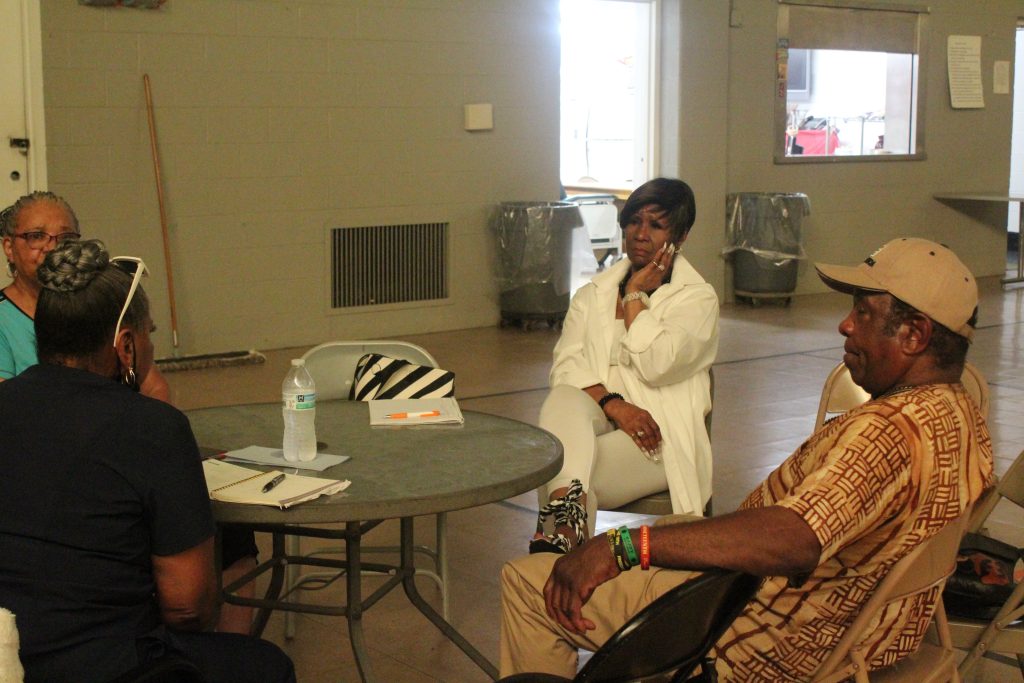 Image: Sylvester "Sunshine" Lee sits at a Rec Center table with three women. The group is holding a planning meeting for a community women’s clothing giveaway to be held at SCAC, 2024. Photo by Treasure Shields Redmond.