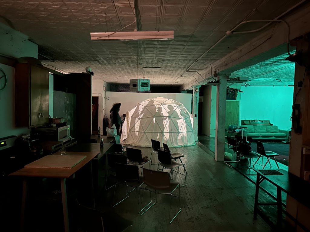 Image: A geometric dome draped in a white cloth sits in the EXP/VOX performance space. Silhouettes of audience members are visible in inside the dome. Outside of the dome are chairs in a high-ceilinged space which includes a kitchen space ot the left and a green lit couch to the right. Photo by Michael Wellvang. Image courtesy of Anna Johnson.