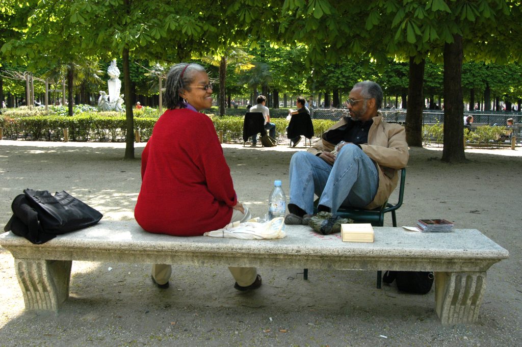 Image: A man and a woman sit on a stone bench in a tree lined park. A fountain sits back of frame. Beverly and William Cottman in Paris, early 2000s. Photo courtesy of writer.