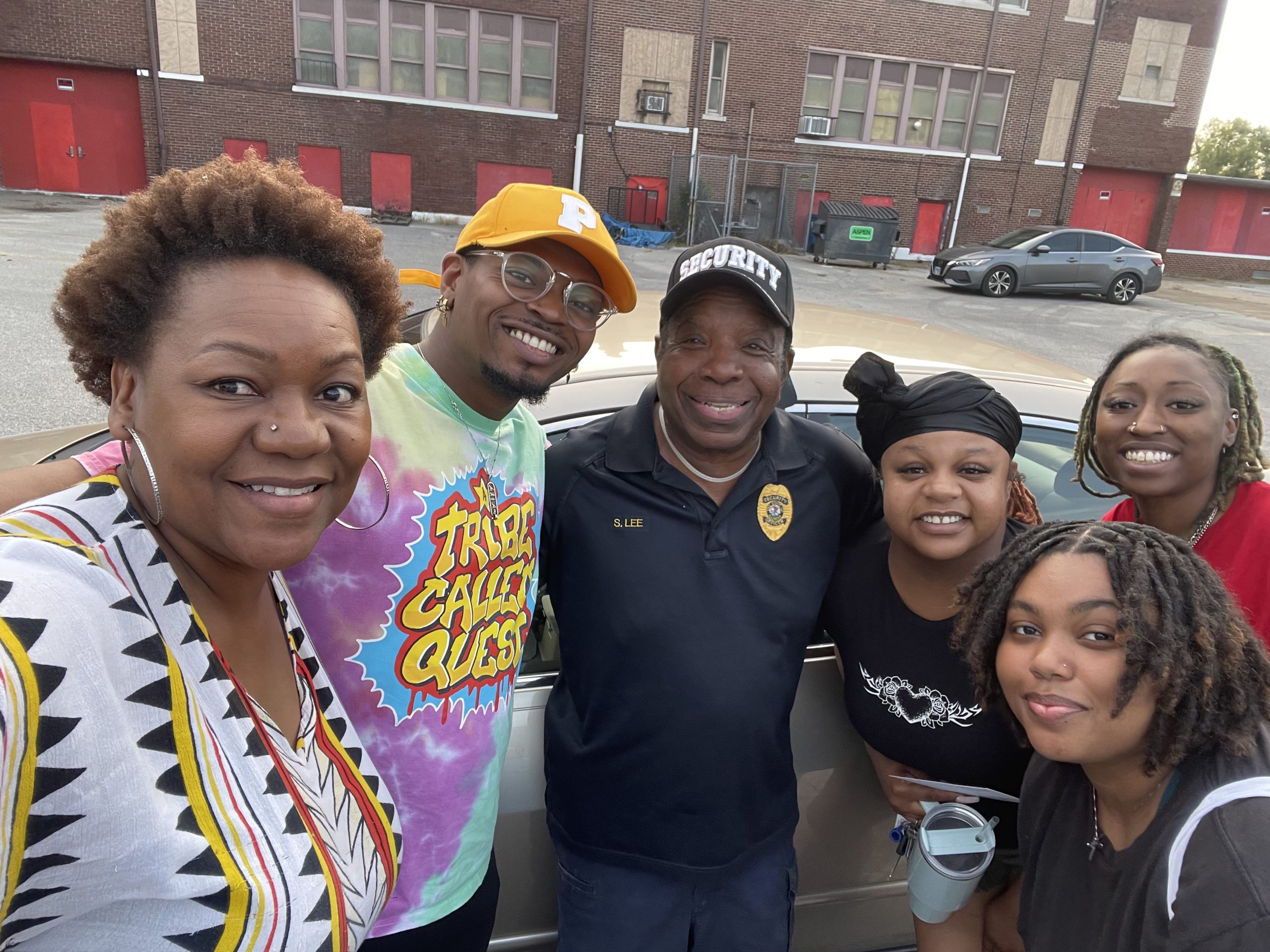 Image: Five smiling people stand center frame in front of a large building. Sunshine and members of The East Side Arts Collective outside of the Sunshine Cultural Arts Center, 2024 (From left to right: Treasure Shields Redmond, Bobby “Phree” Williams, Sylvester "Sunshine" Lee, Elizabeth Stallings, Allena Brazier, and Dominique Shelton)