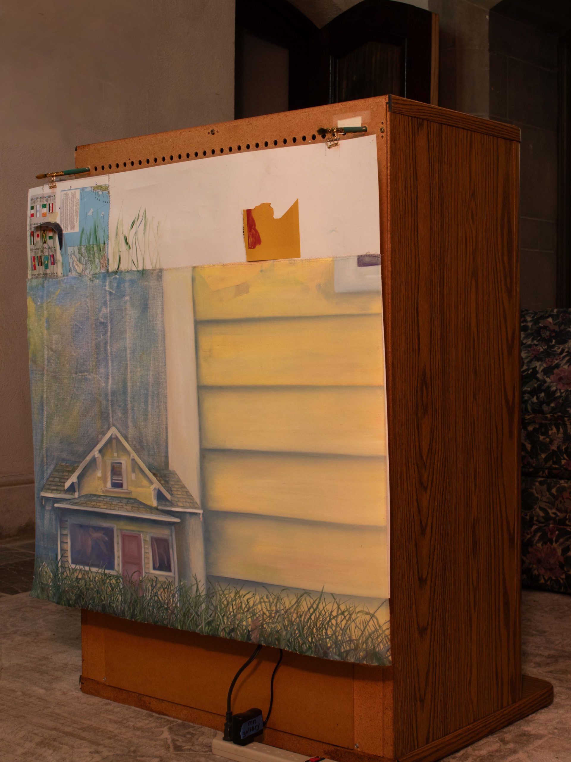 Image: Installation view of the North Narthex (the ground floor). A painted surface is hung on the back of the TV cabinet. On the top-left corner of the surface is a corner of presumably a world map. On the majority of the surface, a small, a medium, and a big house are painted from left to right. On the bottom there is a stripe of grass. The small house is almost covered by the grass and the big house is so big that most of it is cropped out. Below the painted surface, some power cords are connected to a power strip sitting on a greyish carpet with geometric pattern. Photo by Qiuchen Wu.