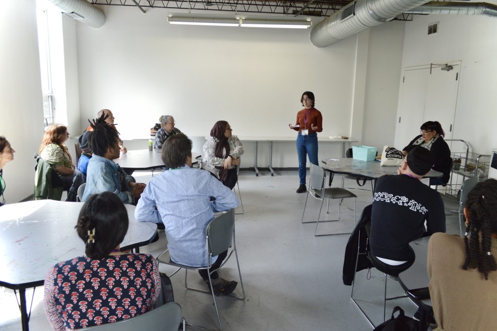 Image: A grey conference room with some daylight filtering in. About fifteen people are seated around at small tables, all looking towards a person standing near the back wall of the room, wearing blue pants and a rust-colored sweater. Their hands are held palm up in front of them. To their left is a double door. Photo by Luz Magdaleno Flores.