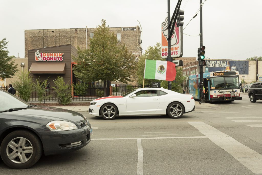 Imagen: Un carro blanco con bandera sobre su cofre y otra bandera por arriba pasa por la intersección de la Kedzie y Cermak Ave., en Chicago. Foto x Cobalto es azul, 2023.