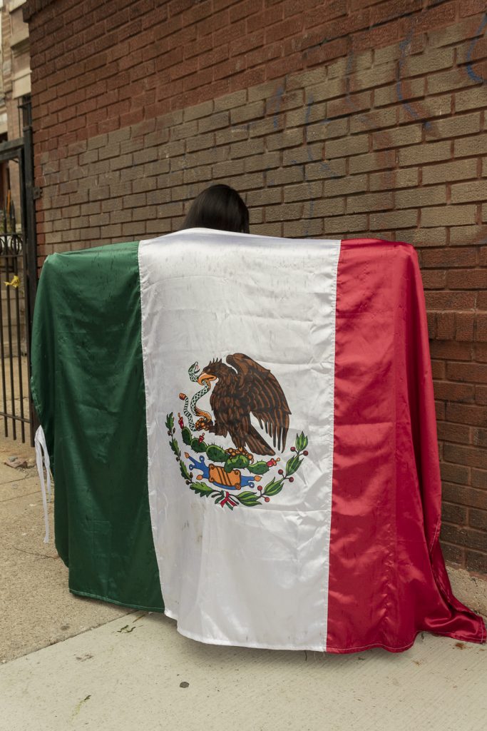 Imagen: Una persona en la calle con una bandera grande en su espalda, su cuerpo se esconde detrás de la bandera y no vemos su cara, solo su pelo cafe. Foto x Cobalto es azul, 2023.