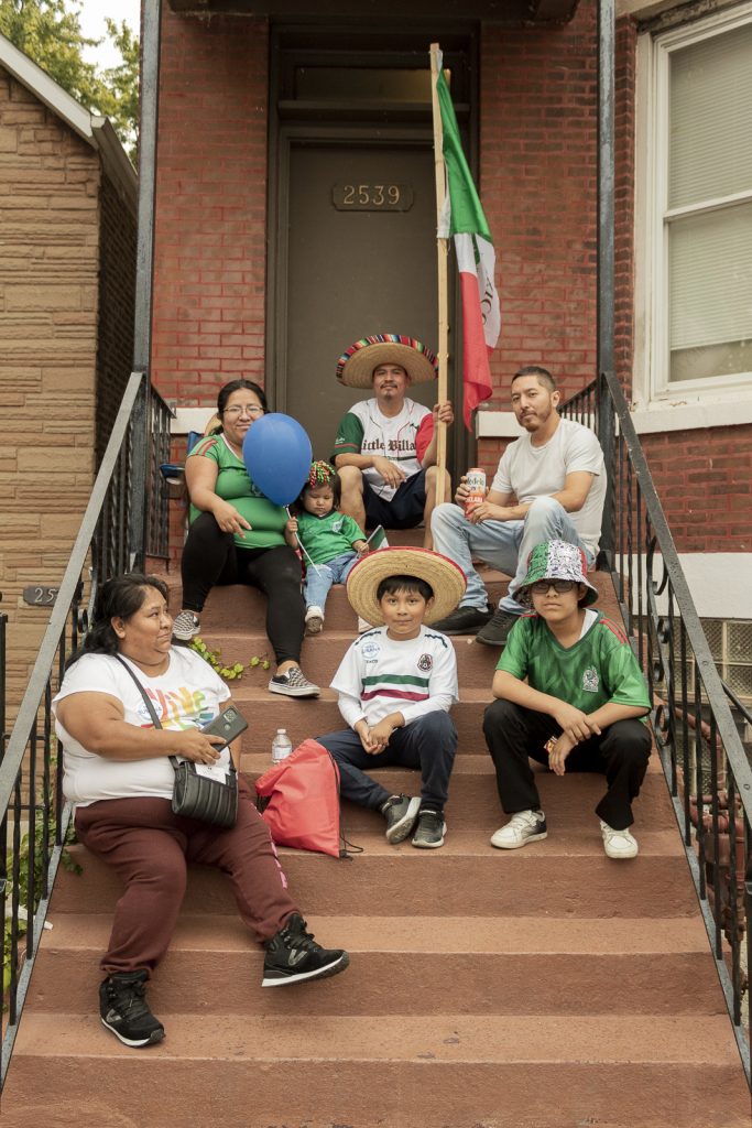 Imagen: Una familia sentada en los escalones del frente de una casa en el bloque 25 de Chicago. Cuatro adultos y tres niños estan vestidos en colores verdes, rojo, y blanco; como la bandera Mexicana. Foto x Cobalto es azul, 2023.