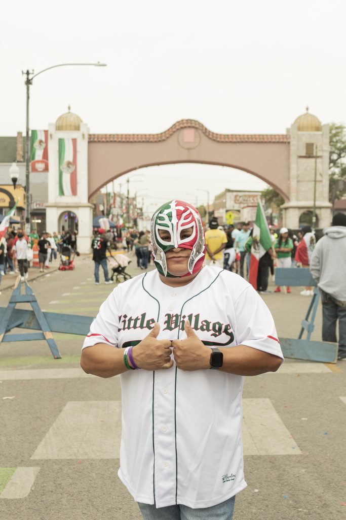 Imagen: Un hombre con mascara de luchador Mexicano y una playera de béisbol con las letras "Little Village" escritas por enfrente posado debajo la arca de la calle 26 en Chicago. Foto x Cobalto es azul, 2023.