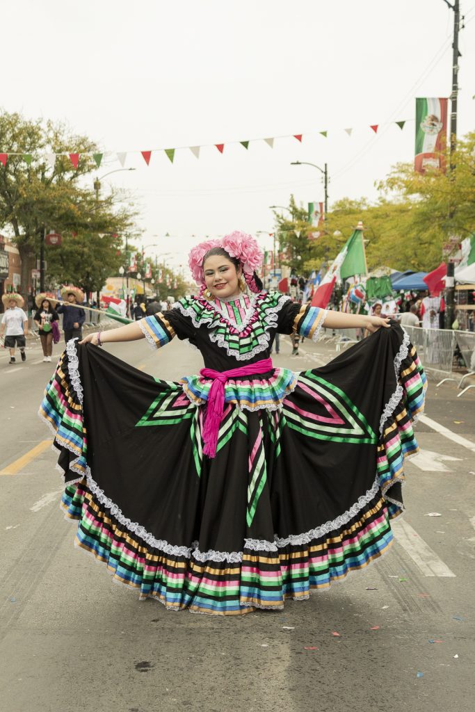 Imagen: Una muchacha con vestido folclórico extendiendo su falda y sonriendo en el desfile de la 26 en Chicago. Foto x Cobalto es azul, 2023.