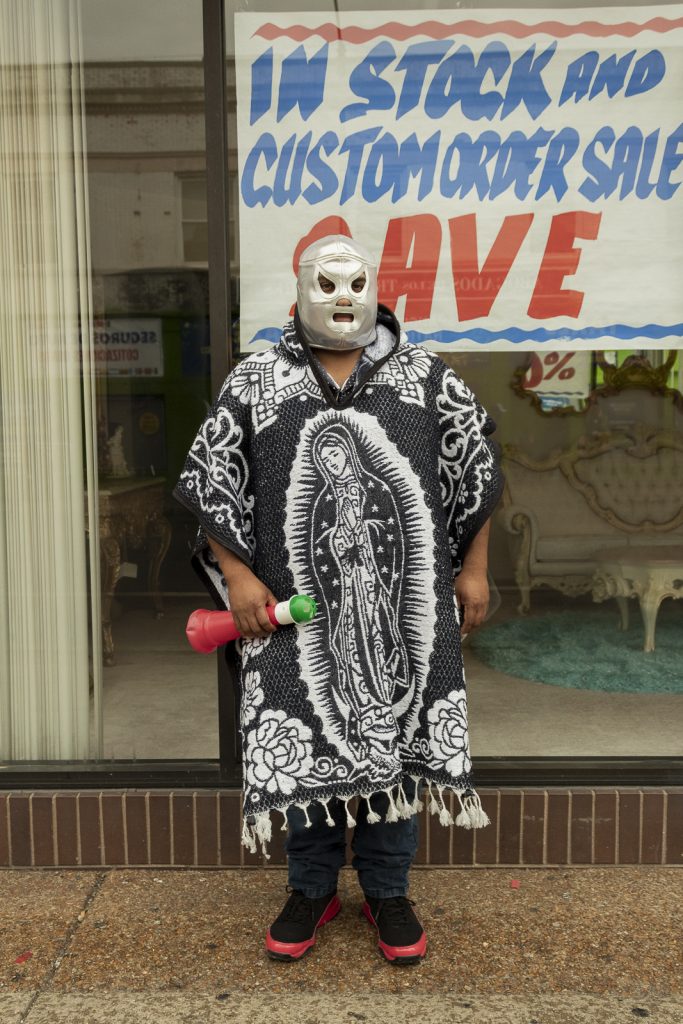 Imagen: Una persona con mascara de luchador y un zarape de la virgen de guadalupe detiene un claxon y posa para la foto en frente de la tienda de muebles en la calle 26 en Chicago. Foto x Cobalto es azul, 2023.